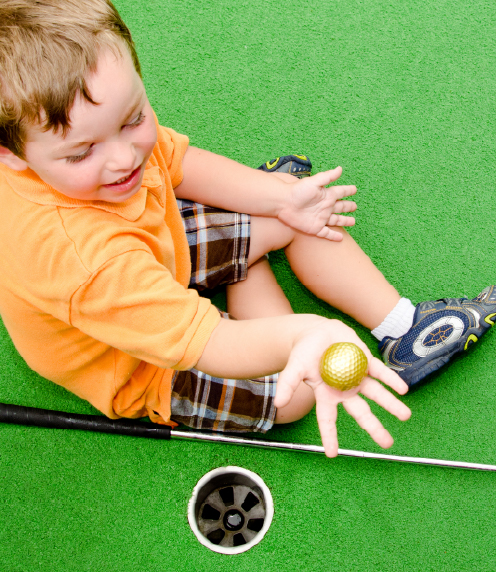 Little boy sitting on astroturf, holding golf ball with golf club next to him