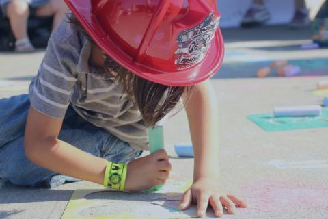 Little girl in fireman's helmet, drawing on sidewalk with chalk