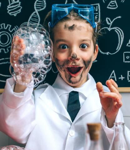 Young boy in lab coat with bubbles in one hand and blackboard of science images behind him
