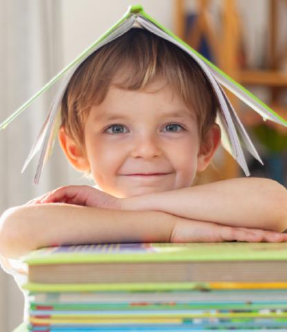 A boy resting his chin on crossed arm set atop a pile of books while wearing an open book as a hat