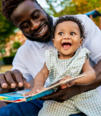 Image: Father and baby daughter smiling while looking at a board book.