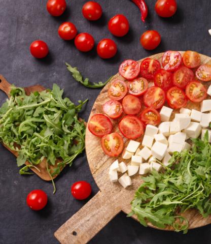 Cutting board with cut up tomatoes, cheese, and greens; surrounded by more of the same on black background