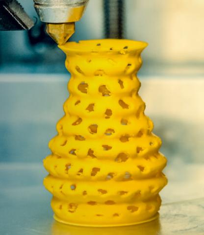 Yellow vase-like mesh form being printed by 3D printer, nozzle in view