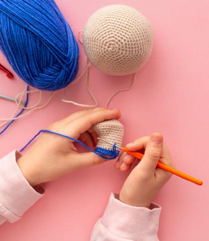 Close up of child's hands crocheting with blue and beige yarn on pink background