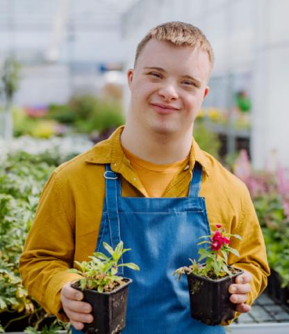 Young man working in garden center, holding up two flowers and smiling proudly