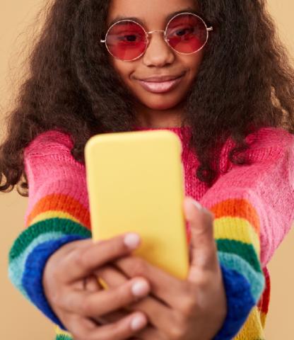 Girl in rainbow colored sweater with rose-tinted glasses holding her smart phone out for a selfie