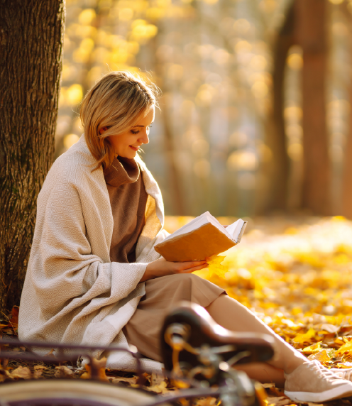 Woman wrapped in a blanket reading book outdoors with autumn leaves