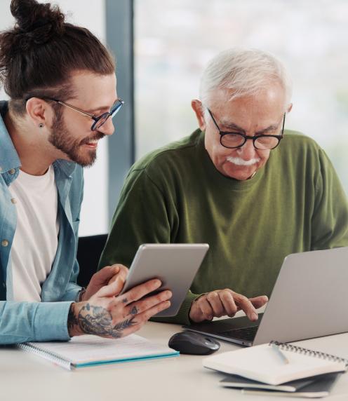 Younger man using a table, sitting next to a senior man on a laptop