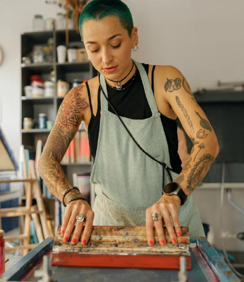 Tattooed woman in workshop apron pulling a squeegee to create a screen print